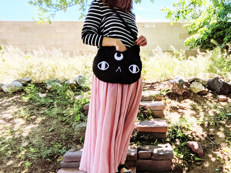 May include: A black crocheted cat-shaped shoulder bag with white eyes and a crescent moon on the forehead. The bag is worn by a person wearing a striped shirt and a pink skirt.