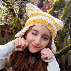 May include: A person wearing a yellow and white striped crocheted cat ear hat with pink ear accents. The hat has a wide brim and is made of soft yarn.