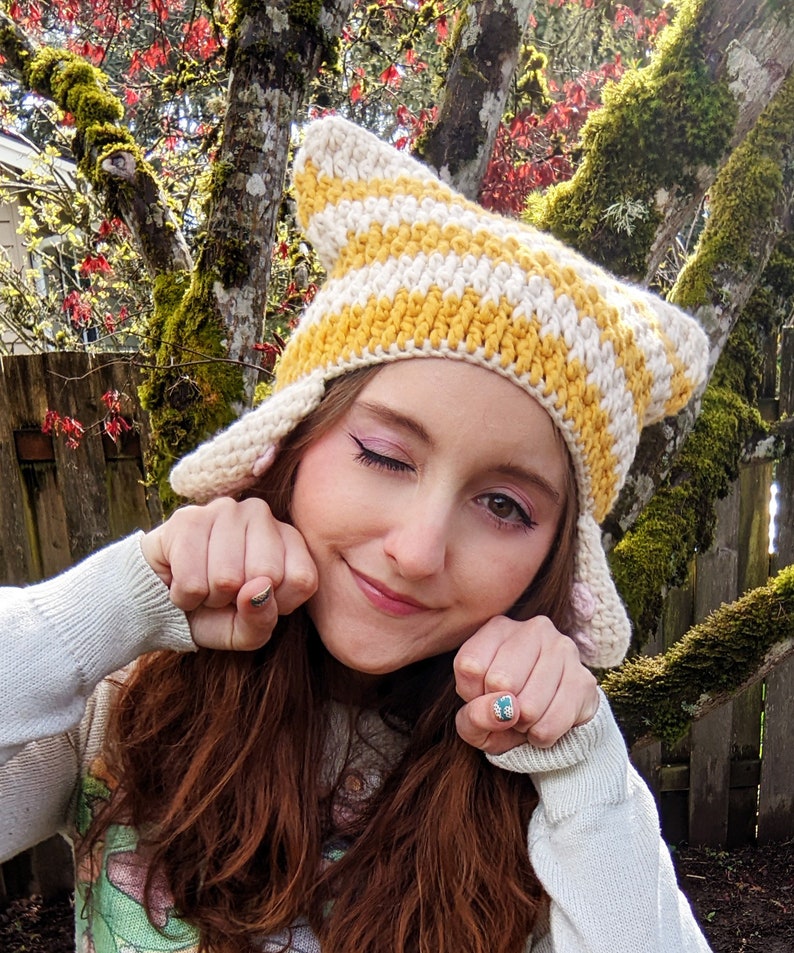 May include: A person wearing a yellow and white striped crocheted cat ear hat with pink ear accents. The hat has a wide brim and is made of soft yarn.