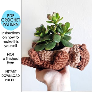 Puede incluir: Patrón de crochet para una maceta de crochet marrón con forma de animal durmiendo. La maceta contiene una pequeña planta suculenta verde.