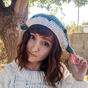 Pu&ograve; includere: Una donna indossa un cappello all'uncinetto bianco e turchese con un design a forma di delfino. Il cappello ha un bordo bianco e un corpo turchese con un motivo all'uncinetto bianco. La donna indossa un maglione bianco.