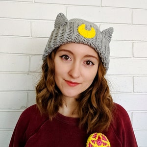 May include: A gray crocheted beanie with cat ears and a yellow crescent moon design. The beanie is worn by a person with long brown hair.