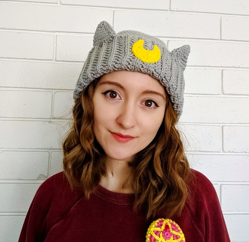 May include: A gray crocheted beanie with cat ears and a yellow crescent moon design. The beanie is worn by a person with long brown hair.