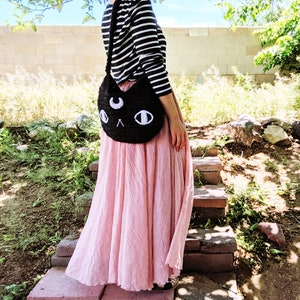 May include: A black crocheted cat-shaped shoulder bag with white eyes and a crescent moon on the front. The bag is worn by a person wearing a striped shirt and a pink skirt.