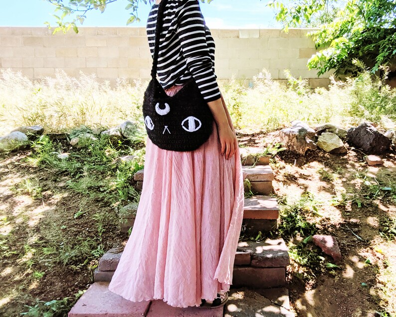 May include: A black crocheted cat-shaped shoulder bag with white eyes and a crescent moon on the front. The bag is worn by a person wearing a striped shirt and a pink skirt.