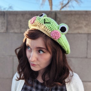 May include: A light green crocheted frog beret with pink and black accents. The frog hat features large, round eyes and a smiling mouth. The beret is a unique, handmade accessory, perfect for adding a whimsical touch to any outfit.