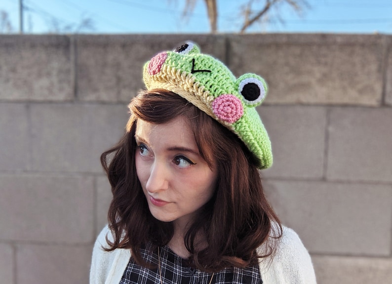 May include: A light green crocheted frog beret with pink and black accents. The frog hat features large, round eyes and a smiling mouth. The beret is a unique, handmade accessory, perfect for adding a whimsical touch to any outfit.