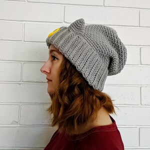 May include: A gray knitted beanie with cat ears and a yellow embroidered design. The beanie is slouchy and has a relaxed fit.