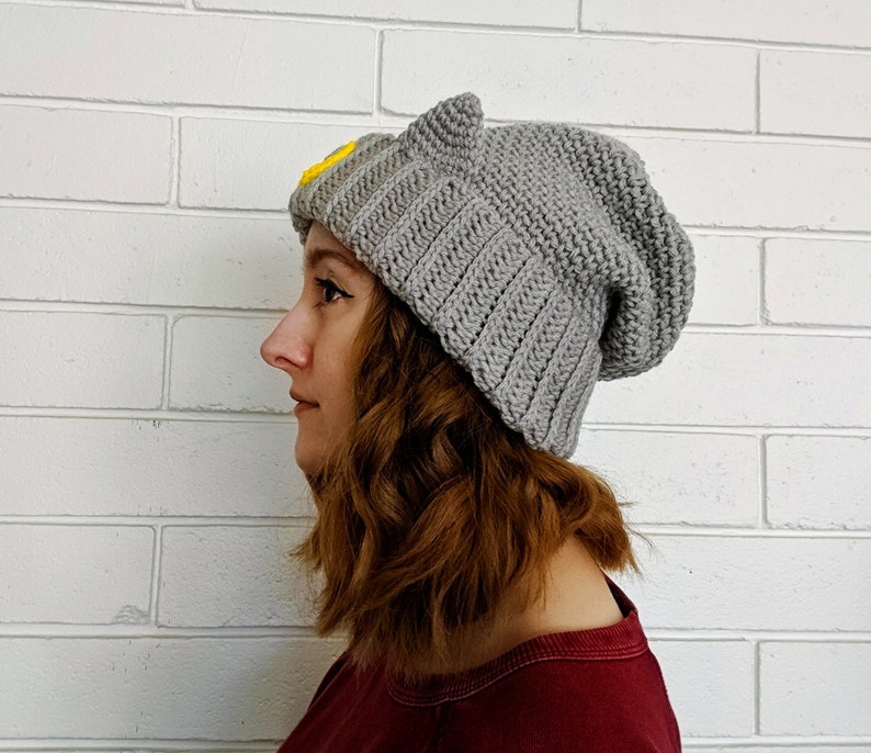May include: A gray knitted beanie with cat ears and a yellow embroidered design. The beanie is slouchy and has a relaxed fit.