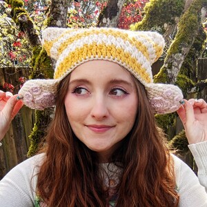 May include: A crocheted hat with yellow and white stripes and pink flower-shaped ear flaps. The hat is being worn by a person with long brown hair.