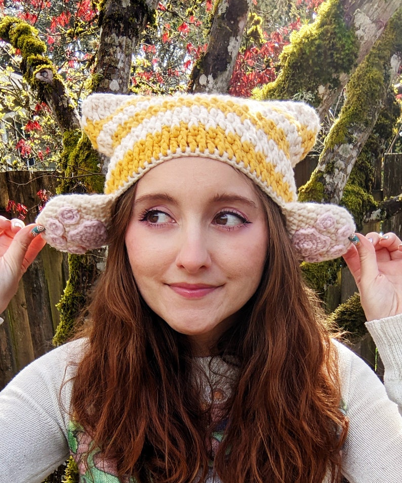 May include: A crocheted hat with yellow and white stripes and pink flower-shaped ear flaps. The hat is being worn by a person with long brown hair.