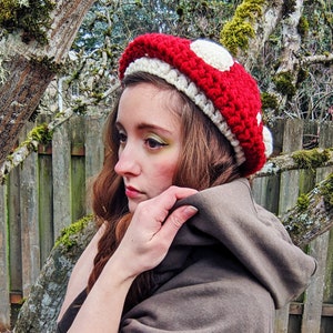 Peut inclure: Une femme portant un chapeau en crochet en forme de champignon rouge et blanc avec une veste &agrave; capuche marron.
