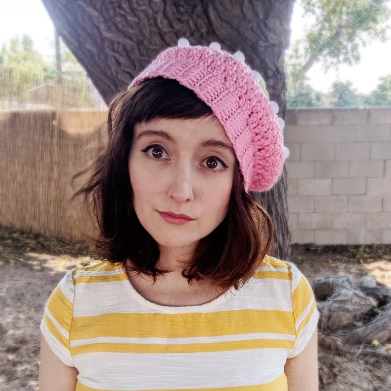 May include: A woman wearing a pink crocheted beret with white pom-poms and a yellow and white striped shirt.