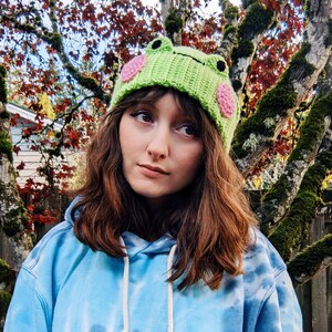 May include: A person wearing a green and pink crocheted frog hat with a blue tie-dye hoodie.