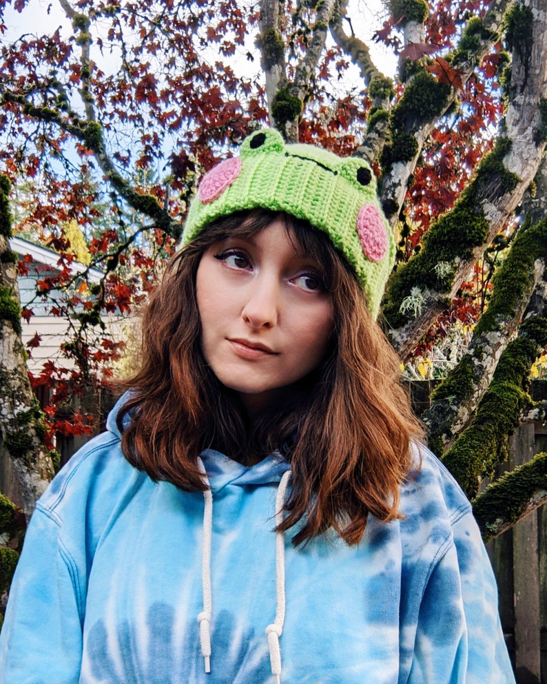 May include: A person wearing a green and pink crocheted frog hat with a blue tie-dye hoodie.
