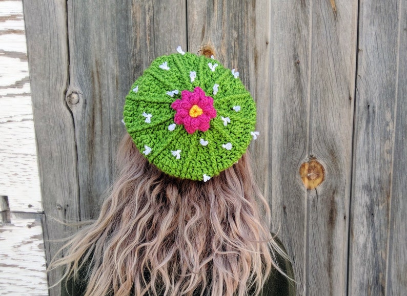 May include: A green crocheted hat shaped like a cactus with a pink and yellow flower in the center.