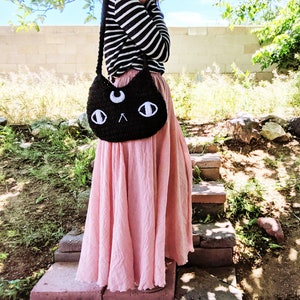 May include: A black crocheted cat-shaped bag with white eyes and a crescent moon on the front. The bag is worn over the shoulder of a person wearing a striped shirt and a pink skirt.