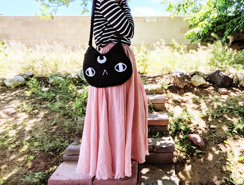 May include: A black crocheted cat-shaped bag with white eyes and a crescent moon on the front. The bag is worn over the shoulder of a person wearing a striped shirt and a pink skirt.