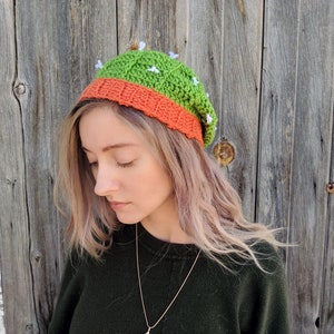 May include: A green and orange crocheted beanie hat with white flower accents. The hat is worn by a person with long blonde hair.