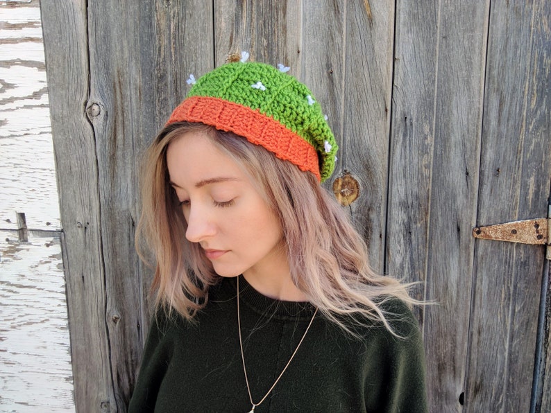 May include: A green and orange crocheted beanie hat with white flower accents. The hat is worn by a person with long blonde hair.