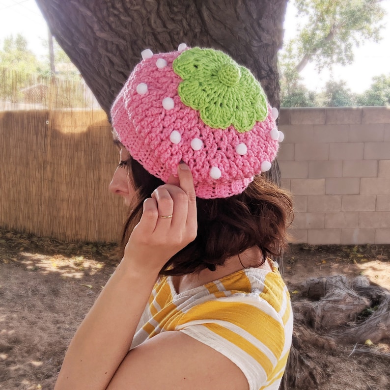 May include: A pink and green crocheted hat shaped like a strawberry. The hat has white pom-poms around the edge.