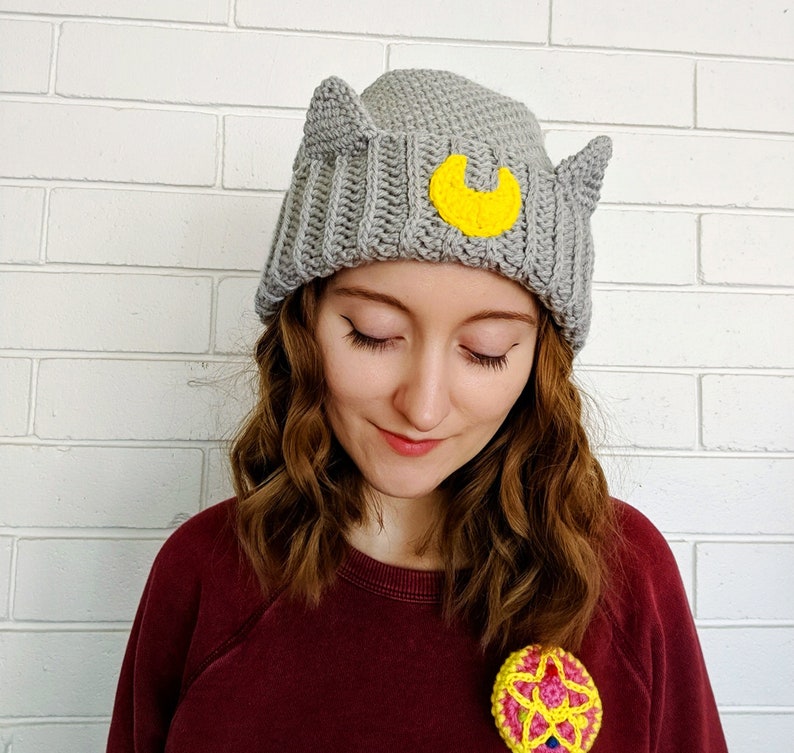 May include: A gray crocheted beanie with cat ears and a yellow crescent moon design. The beanie is worn by a person with long brown hair.