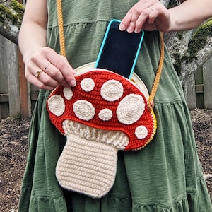 Op de afbeelding: Een gehaakte tas in de vorm van een paddenstoel met een rode bovenkant en witte stippen, en een beige onderkant. Een zwarte smartphone is zichtbaar in de tas.