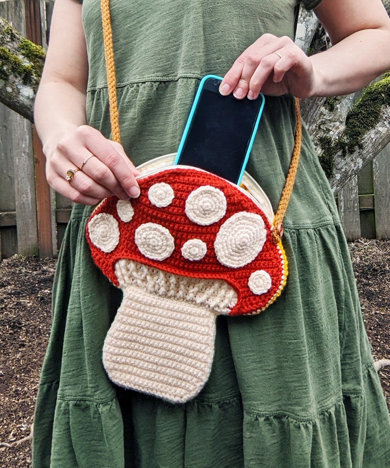 Op de afbeelding: Een gehaakte tas in de vorm van een paddenstoel met een rode bovenkant en witte stippen, en een beige onderkant. Een zwarte smartphone is zichtbaar in de tas.