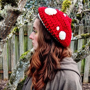 Peut inclure: Un chapeau en crochet rouge en forme de champignon avec des pois blancs. Le chapeau est port&eacute; par une personne aux cheveux longs et bruns.