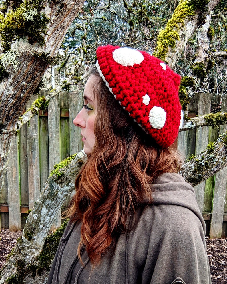 Peut inclure: Un chapeau en crochet rouge en forme de champignon avec des pois blancs. Le chapeau est port&eacute; par une personne aux cheveux longs et bruns.