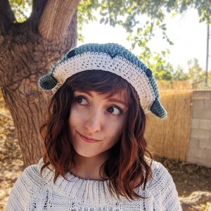 Pu&ograve; includere: Una donna indossa un cappello all'uncinetto blu e bianco. Il cappello ha un design fantasioso con una forma arrotondata e un piccolo orecchio appuntito su ogni lato. La donna ha i capelli lunghi e castani e indossa un maglione bianco.