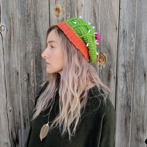 May include: A woman wearing a green and orange crocheted cactus hat with white accents. The hat has a pink flower on the side. She is wearing a dark green sweater and has long blonde hair.