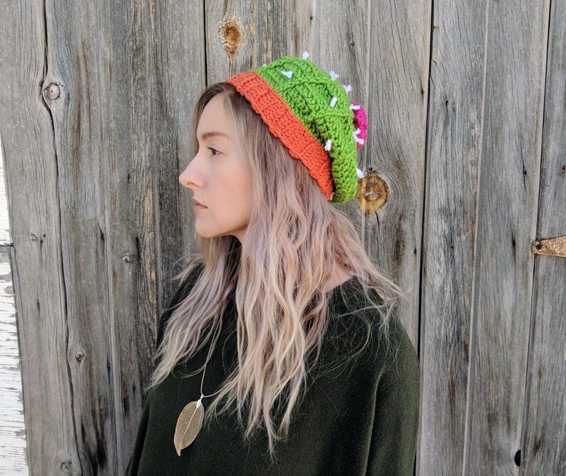 May include: A woman wearing a green and orange crocheted cactus hat with white accents. The hat has a pink flower on the side. She is wearing a dark green sweater and has long blonde hair.
