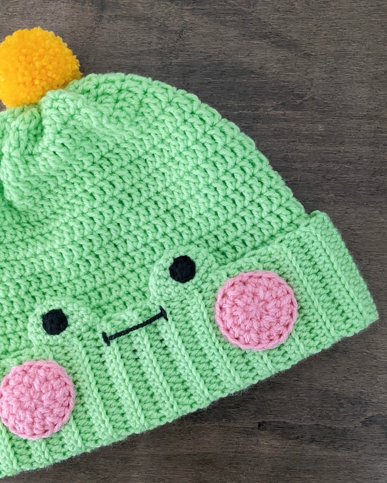 May include: Green crocheted hat with a yellow pom-pom on top and a cute frog face with pink cheeks and black eyes.