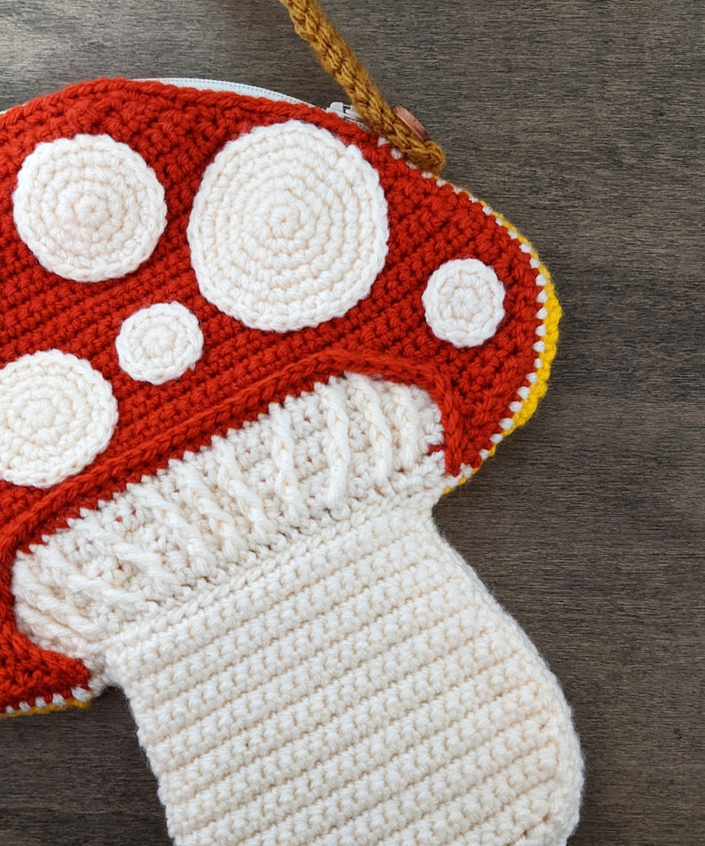 Op de afbeelding: Een gehaakte tas in de vorm van een paddenstoel met een rood en wit bovendeel en een witte steel. De tas heeft een ritssluiting en een gele en bruine band.