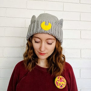 May include: A gray crocheted beanie with cat ears and a yellow crescent moon design. The beanie is worn by a person with long brown hair. The person is wearing a maroon sweater with a yellow and pink crocheted flower brooch.