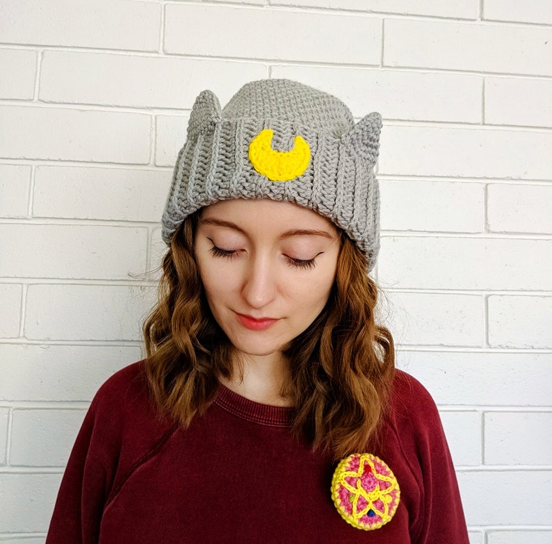 May include: A gray crocheted beanie with cat ears and a yellow crescent moon design. The beanie is worn by a person with long brown hair. The person is wearing a maroon sweater with a yellow and pink crocheted flower brooch.