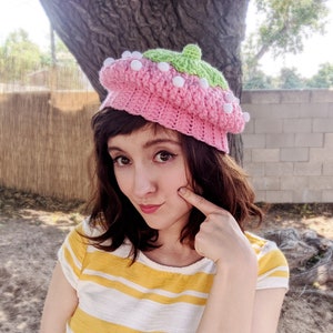 May include: A woman wearing a pink and green crocheted beret with white pom-poms. The beret has a textured design and is decorated with white pom-poms.