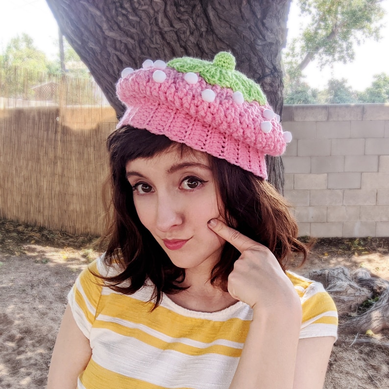 May include: A woman wearing a pink and green crocheted beret with white pom-poms. The beret has a textured design and is decorated with white pom-poms.