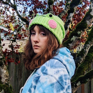 May include: A green crocheted beanie with a frog face and pink spots. The beanie is being worn by a person with long brown hair.