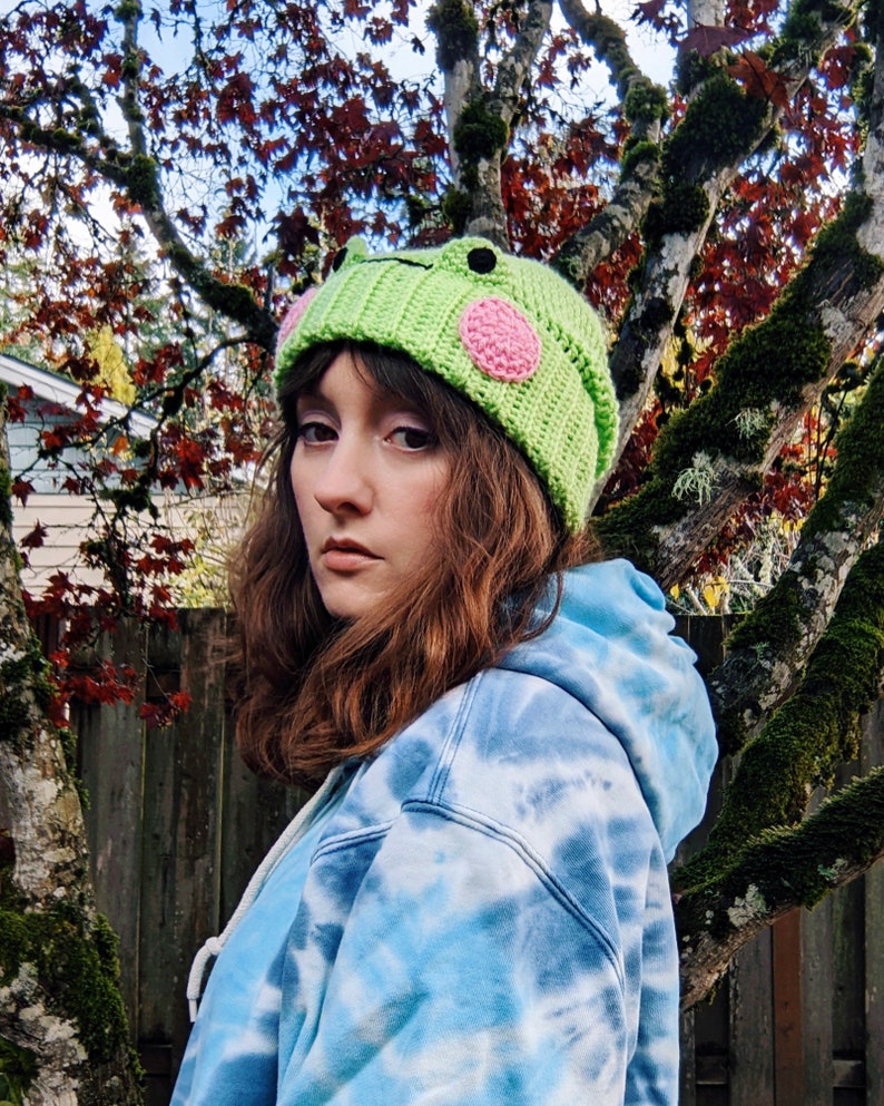 May include: A green crocheted beanie with a frog face and pink spots. The beanie is being worn by a person with long brown hair.