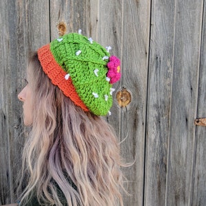 May include: A green and orange crocheted hat shaped like a cactus with a pink flower on top.