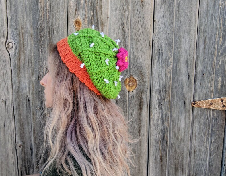 May include: A green and orange crocheted hat shaped like a cactus with a pink flower on top.