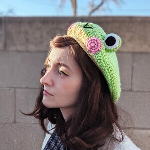May include: A light green crochet frog beret with pink and black accents. The hat features a frog face design with large eyes and a smiling mouth. The beret is a unique and whimsical accessory.