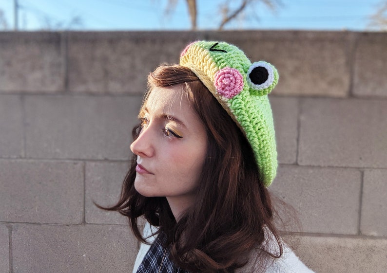 May include: A light green crochet frog beret with pink and black accents. The hat features a frog face design with large eyes and a smiling mouth. The beret is a unique and whimsical accessory.