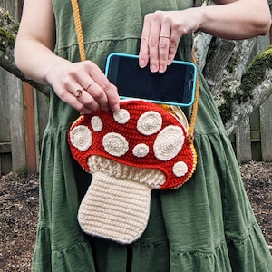 Op de afbeelding: Een gehaakte tas in de vorm van een paddenstoel met een rode bovenkant en witte stippen, en een cr&egrave;mekleurige steel. De tas wordt als schoudertas gedragen en heeft een telefoon erin.