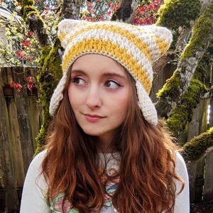May include: A woman with long, auburn hair is wearing a crocheted hat with yellow and white stripes. The hat has ear flaps and is styled like a cat's ears.