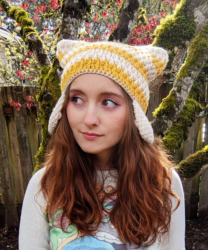 May include: A woman with long, auburn hair is wearing a crocheted hat with yellow and white stripes. The hat has ear flaps and is styled like a cat's ears.