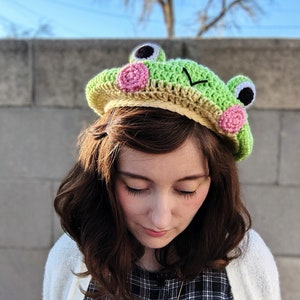 May include: A handmade crochet frog hat in shades of green, tan, and pink. The hat features large, round eyes, pink cheek accents, and a smiling mouth. The hat is designed to be worn on the head.