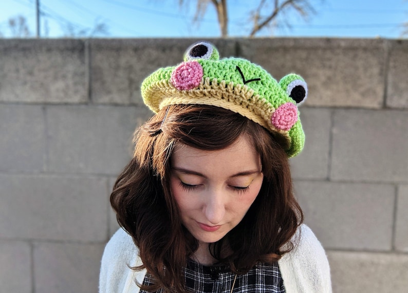May include: A handmade crochet frog hat in shades of green, tan, and pink. The hat features large, round eyes, pink cheek accents, and a smiling mouth. The hat is designed to be worn on the head.